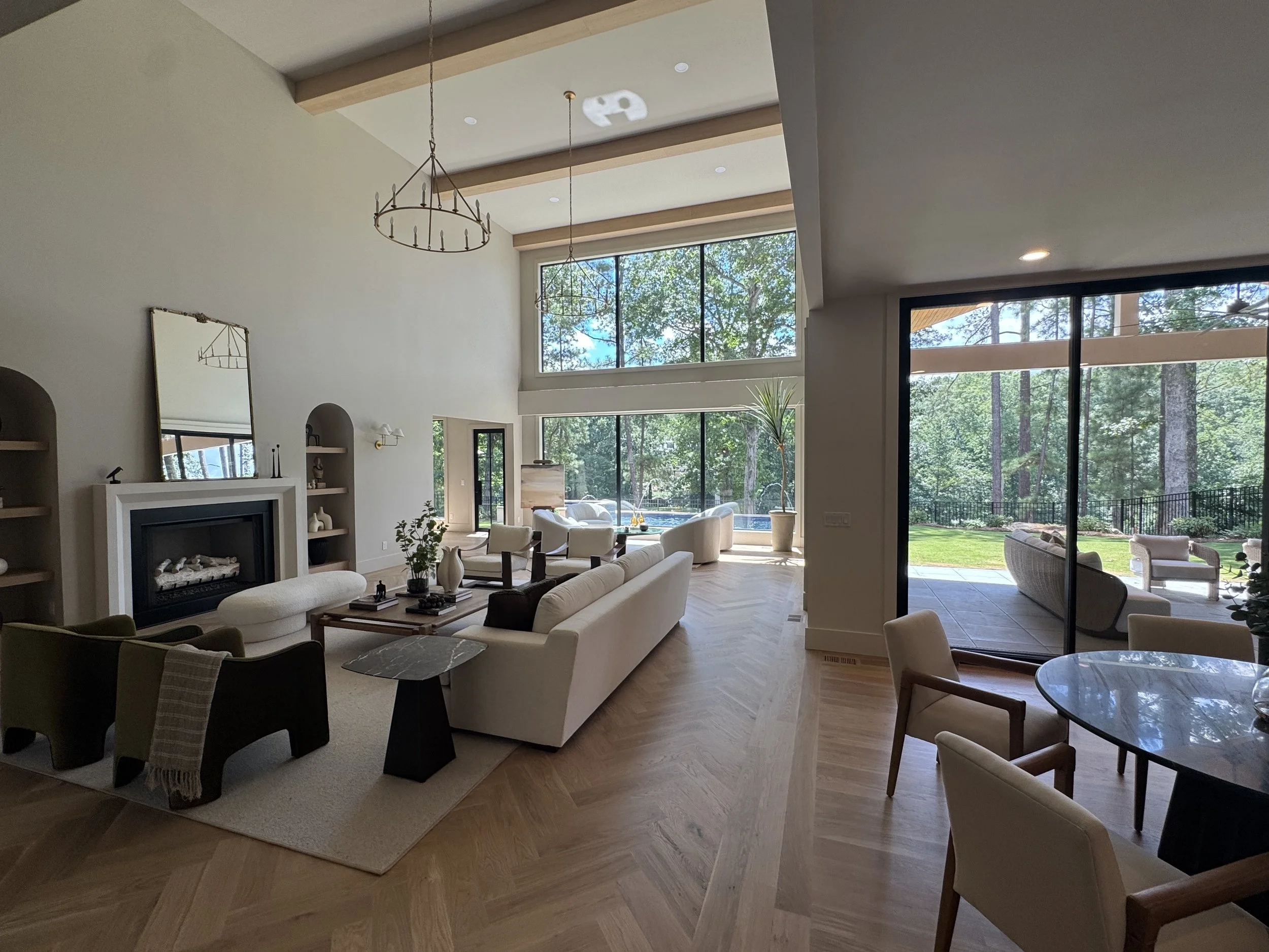 Spacious living room with large floor-to-ceiling windows showing trees outside. Modern furniture including white sofas, chairs, and a marble-topped table. Wooden floors and a high ceiling with wooden beams and chandeliers. Patio area visible through 