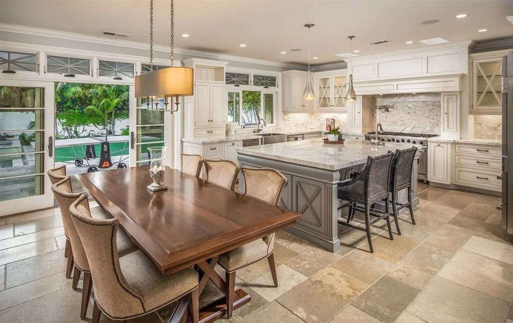 A spacious kitchen and dining area with beige tiled floors, white cabinetry, and a large island with a marble countertop. There are black chairs around the island and a wooden dining table with beige upholstered chairs. In the background, sliding gla
