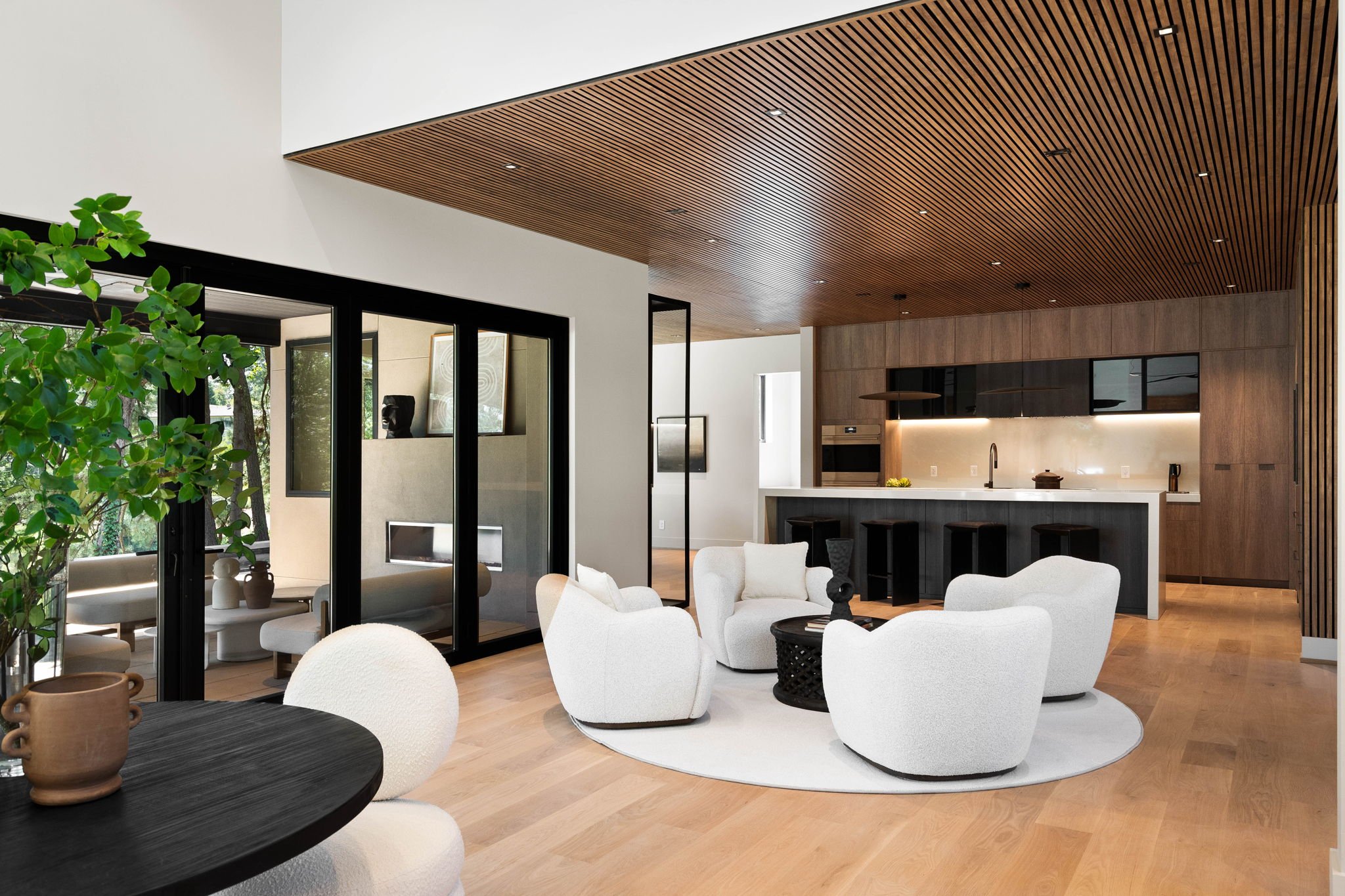 Modern living room with white plush chairs, a black round table, and a large indoor plant in the foreground. Open kitchen area with wood cabinets, black accents, and minimalist decor. Ceiling lined with wooden slats with recessed lighting, and large 