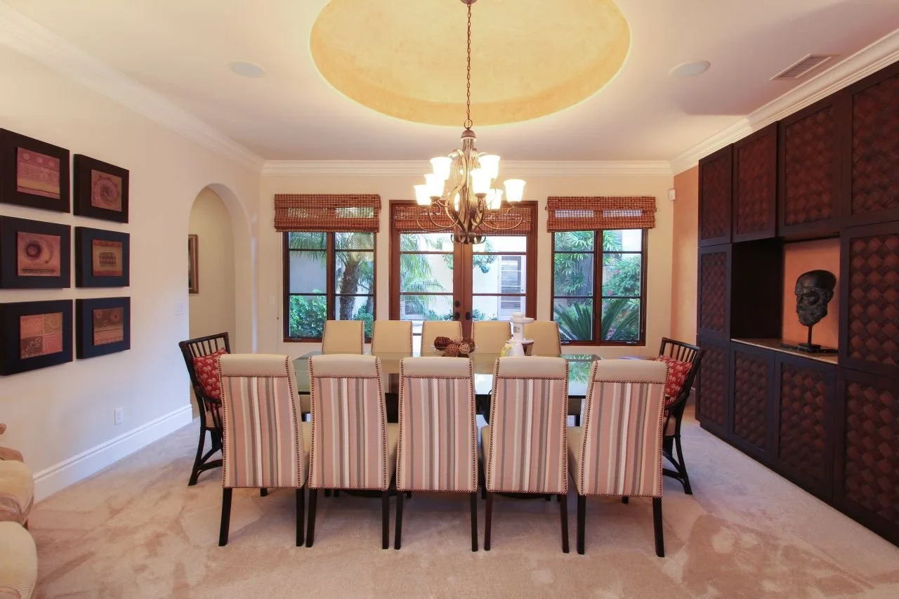 A dining room with a large table surrounded by striped and wooden chairs, a chandelier hanging from a recessed ceiling, windows with bamboo blinds, framed artwork on the wall, and a built-in dark wood cabinet with decorative items.