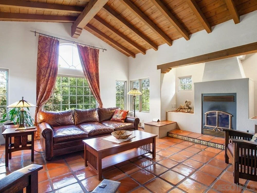 Living room with a large window, wooden ceiling, leather sofa, wooden coffee table, side tables with lamps, tiled floor, and a fireplace with a sculpture on the bench nearby.