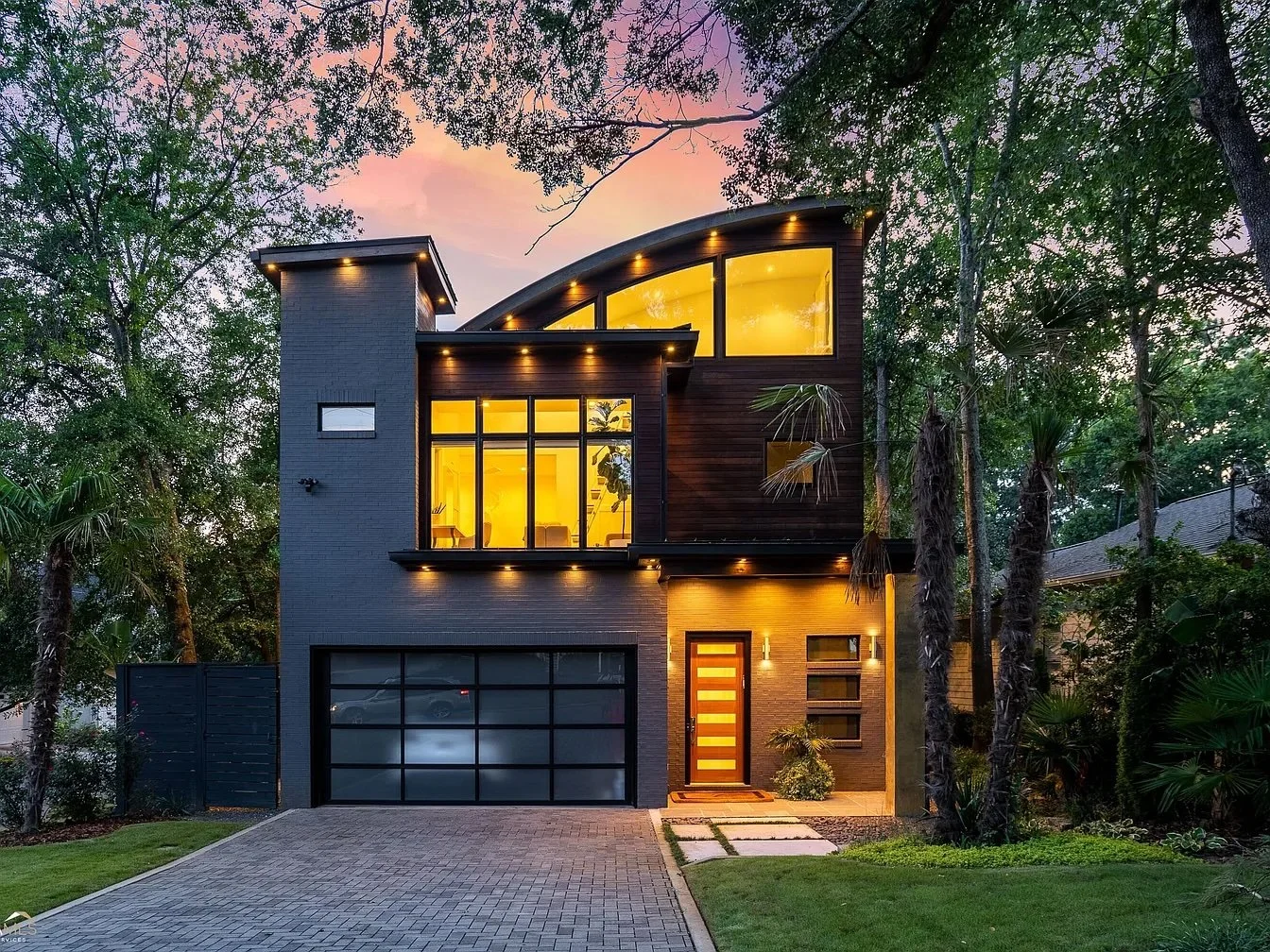 Modern two-story house with illuminated windows, surrounded by trees in the evening, with a paved driveway and neatly maintained lawn.