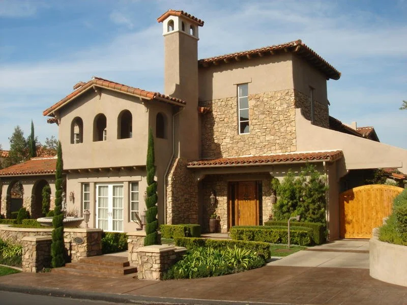 A large, multi-story house with beige stucco walls, stone accents, and a red tile roof, surrounded by well-maintained greenery and a small front yard with a stone pathway.