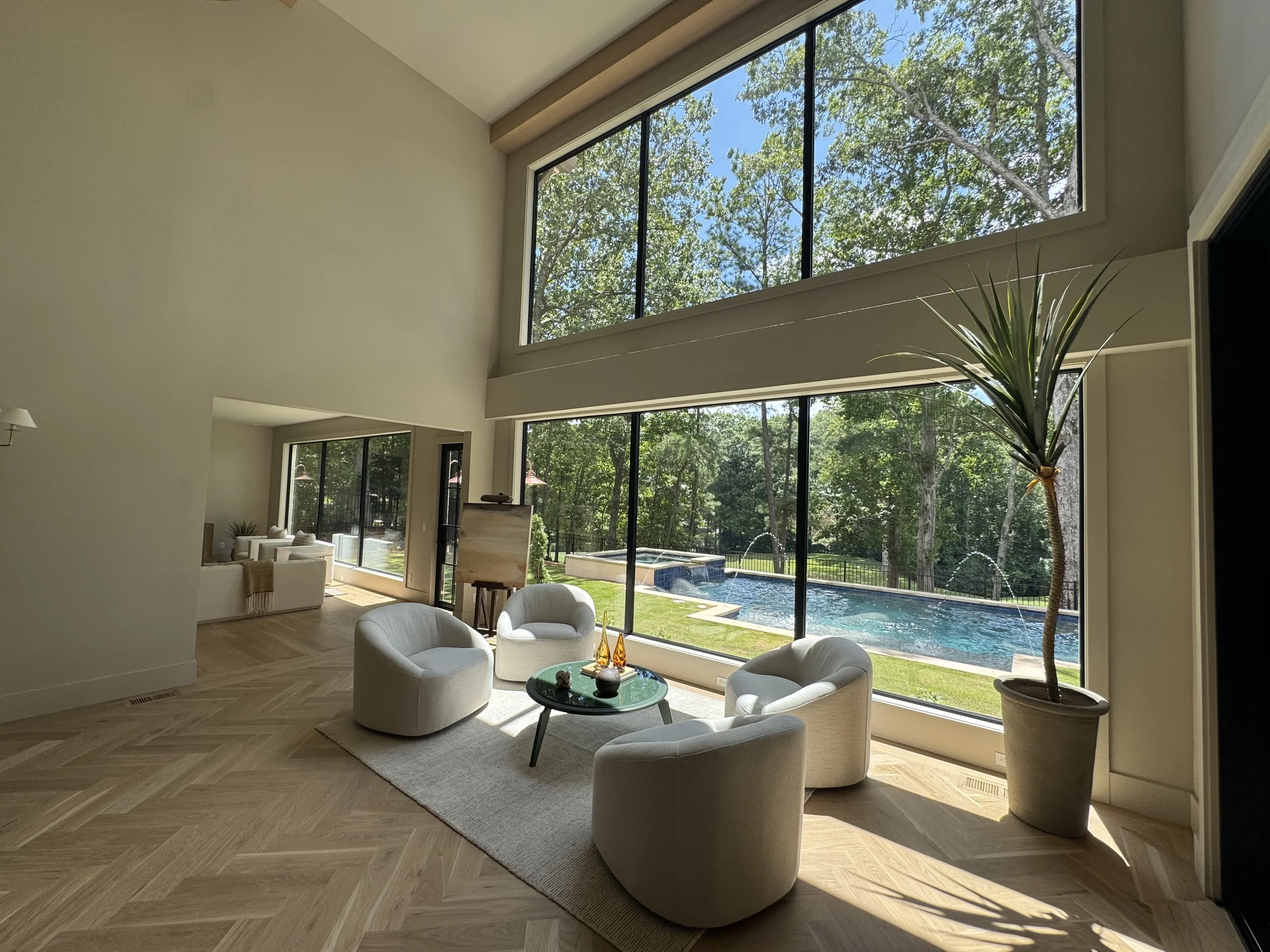 A spacious living room with large floor-to-ceiling windows overlooking a backyard with green trees and a swimming pool. The room features a white seating area, a small black coffee table, a tall potted plant, and light wood flooring.