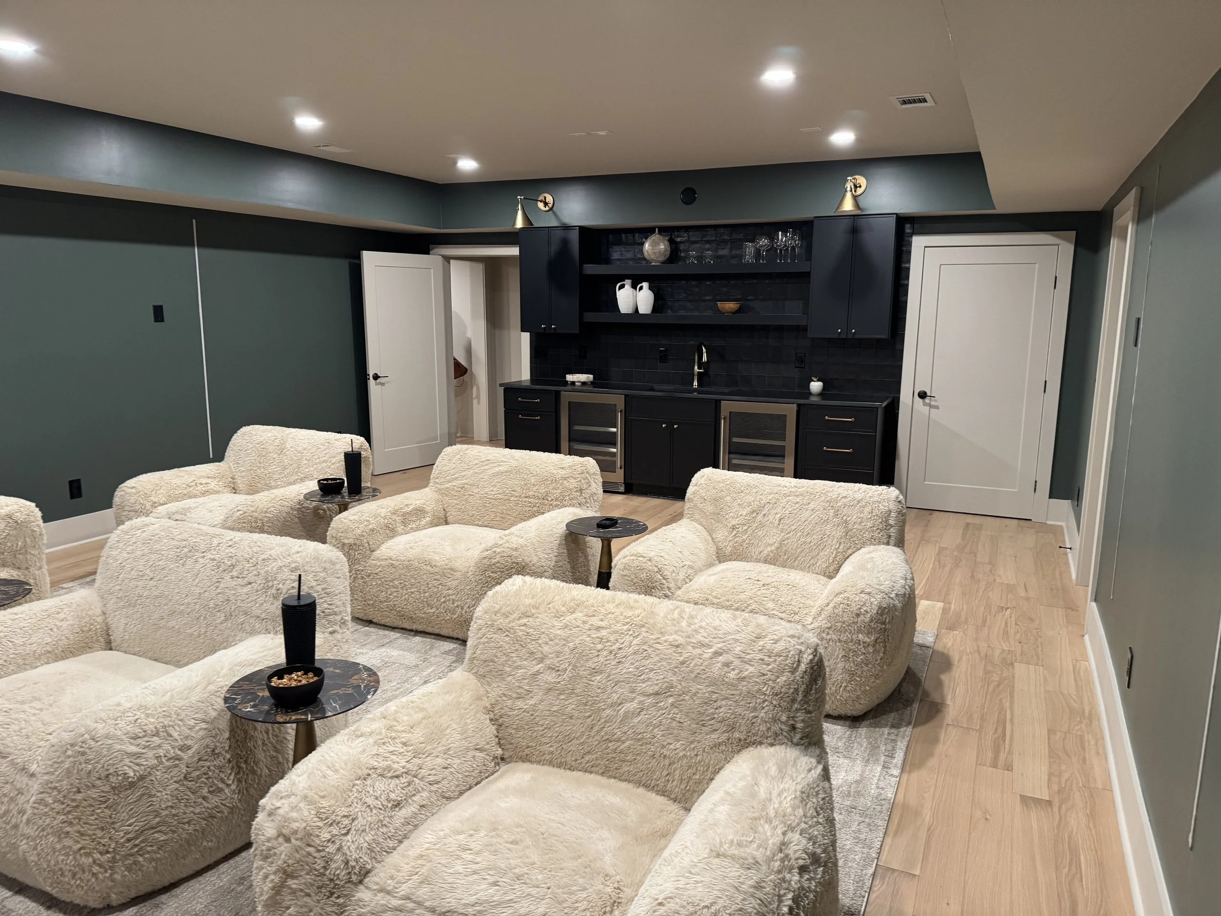 Living room with plush cream chairs, a small marble-top table, a black kitchen cabinet with open shelving, and closed doors, with recessed ceiling lights and wood flooring.