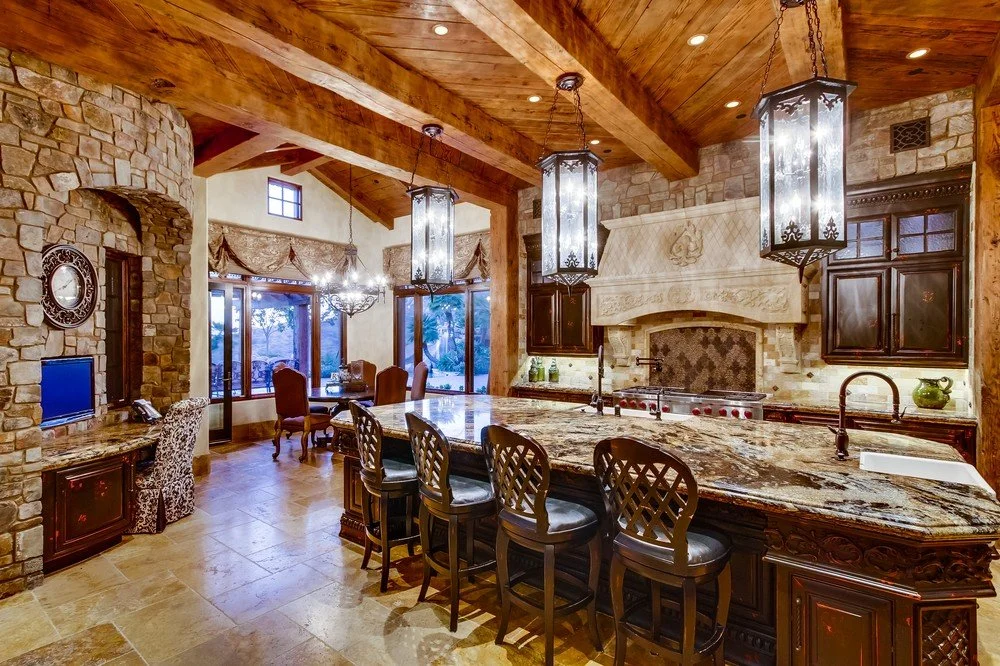 A rustic kitchen with a large granite island, dark wooden cabinetry, stone walls, and wooden beam ceilings. There are hanging lantern-style lights above the island and a windowed dining area in the background.