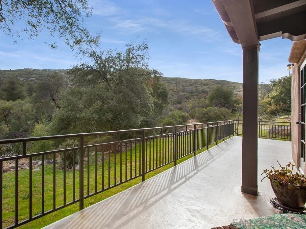 View from a balcony overlooking a grassy yard with trees and hilly landscape in the distance. The balcony has a black metal railing and part of the house's exterior is visible on the right.