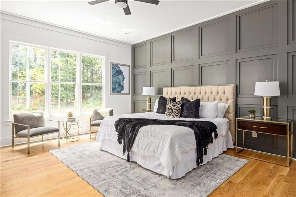 Bright bedroom with large window, gray paneled accent wall, beige tufted headboard, white bedding with black and patterned pillows, black throw blanket, two gray armchairs by window, gold and white side tables, white lamps, light wood flooring, abstr