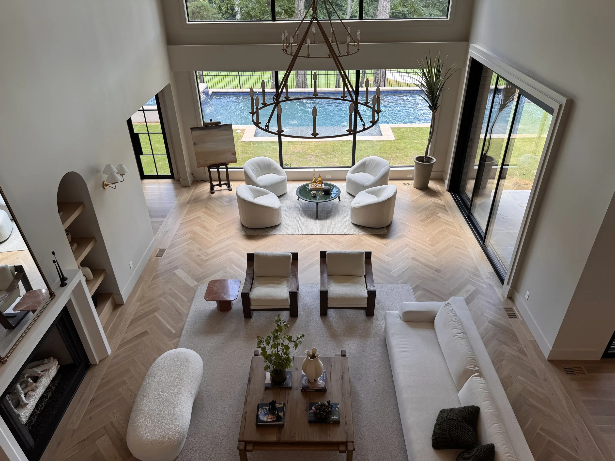 Living room with a high ceiling, a large chandelier, and floor-to-ceiling windows overlooking a backyard with a swimming pool. There are four white armchairs around a small round table, a fireplace on the left, and decor plants.