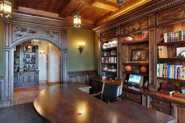 A wood-paneled office with a large curved desk, built-in bookshelves, and decorative woodwork. There are shelves with books and sports memorabilia, a computer on the desk, and a wall-mounted light fixture. An archway leads to another room with a disp