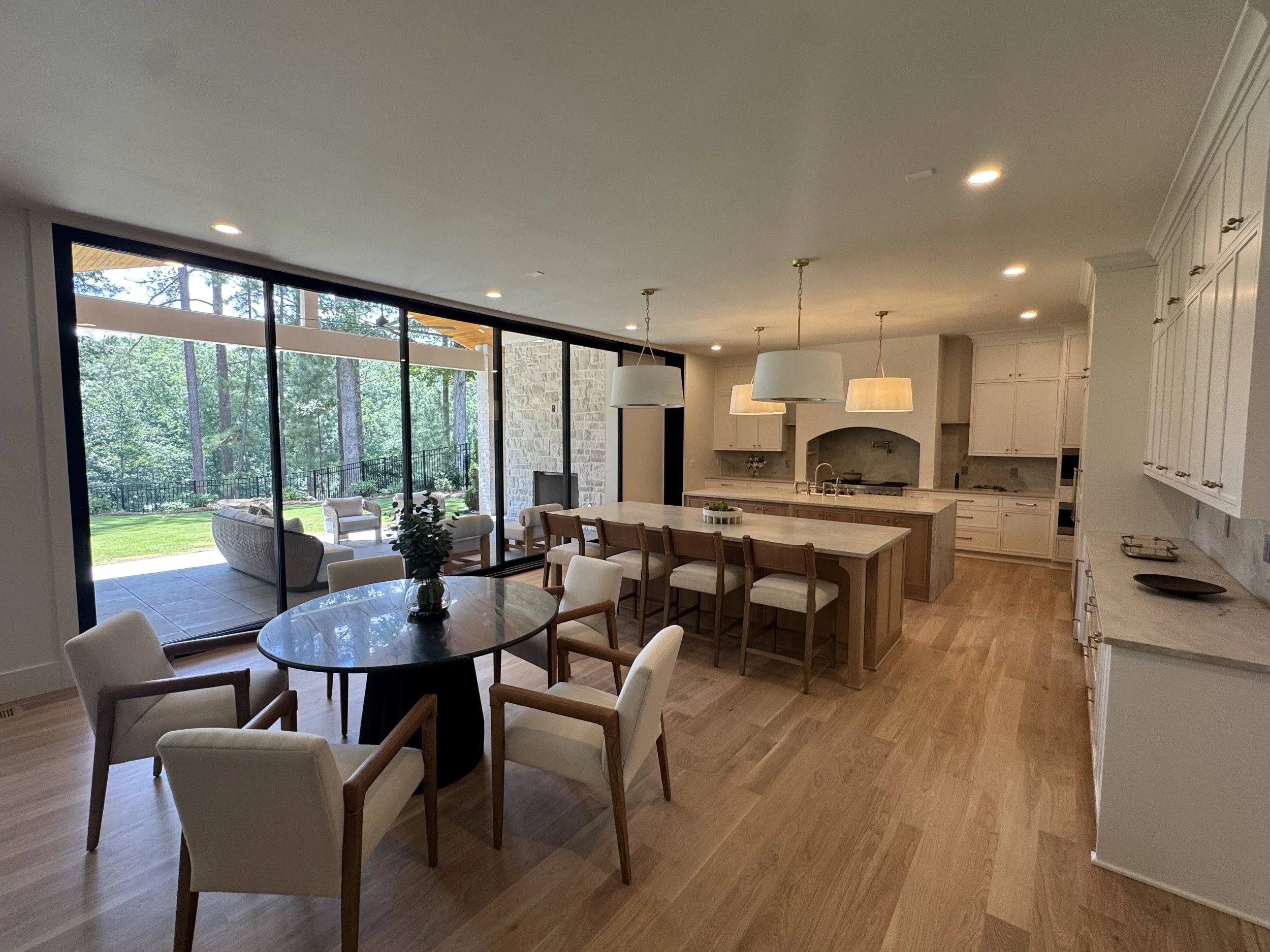 Open-concept living space with large windows showing a wooded backyard, a kitchen with white cabinets and island, and a dining area with a round table and white chairs.