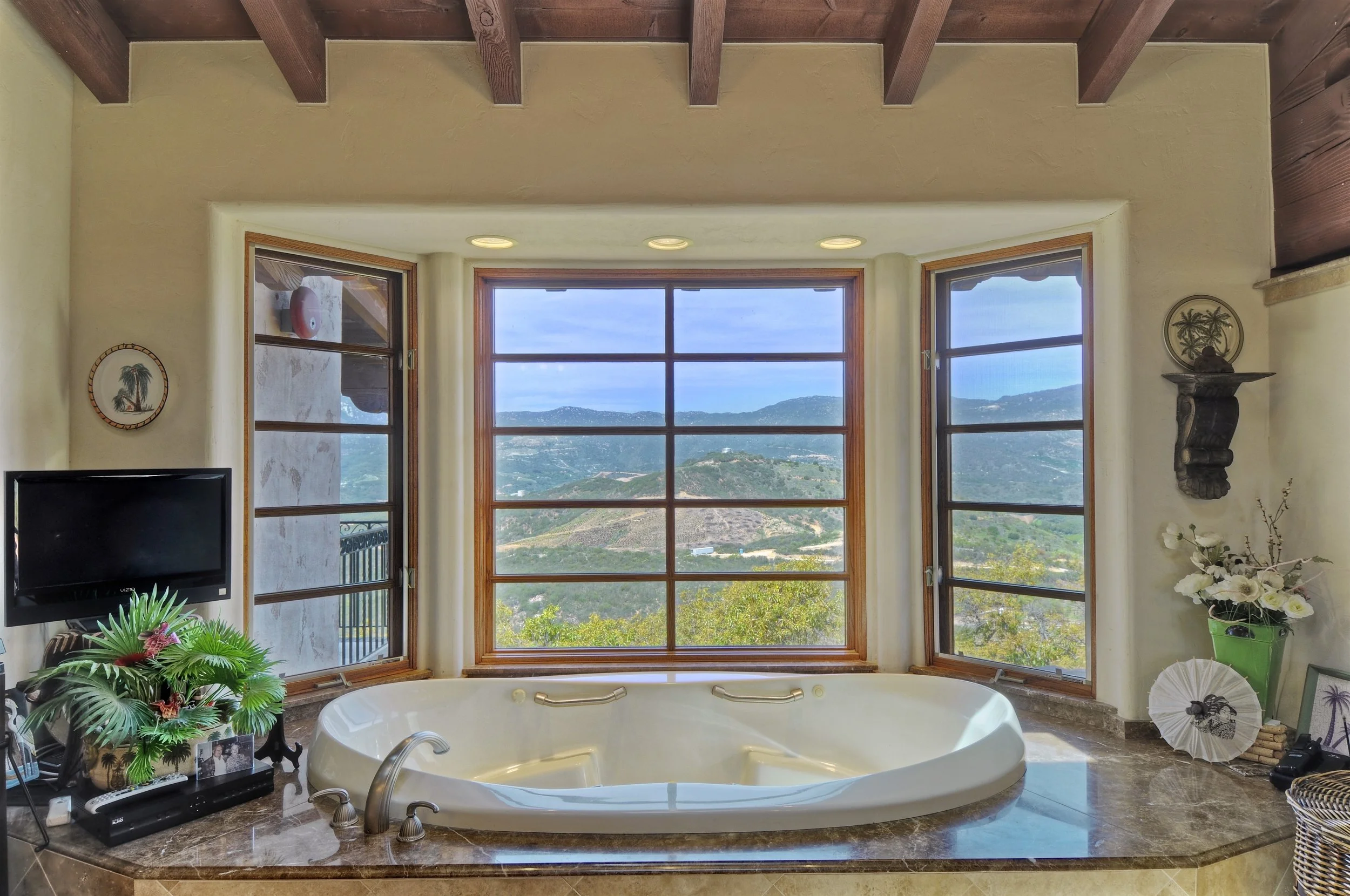 A bathroom with a large bathtub positioned in front of three tall windows overlooking a scenic mountain landscape. The room has beige walls, decorative pieces, a small television, plants, and sculptures. The ceiling features exposed wooden beams.
