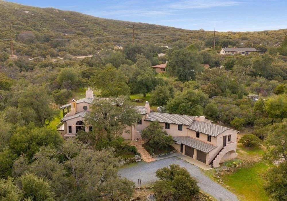 A large, multi-story house surrounded by trees and greenery, with a gravel driveway leading to a garage, located in a hilly area with other houses visible in the background.