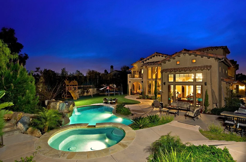 Luxury house with outdoor swimming pool and hot tub during evening, illuminated interior, surrounded by lush greenery and garden furniture.