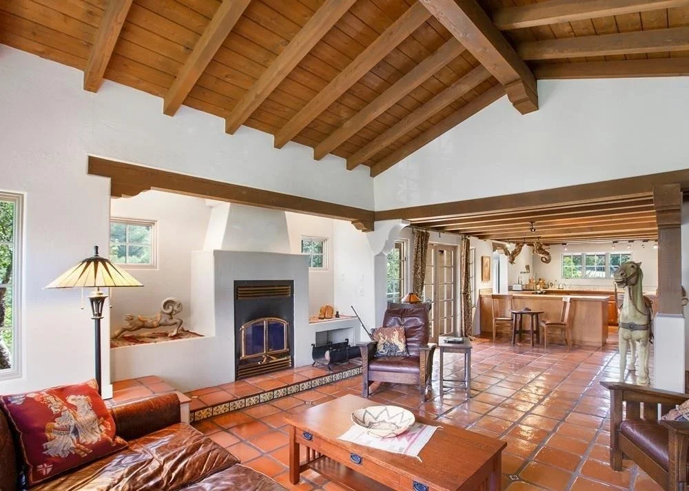Living room with white walls, a fireplace, leather chairs, and a wooden ceiling. The room has terracotta tiled flooring, and the open kitchen area can be seen in the background.