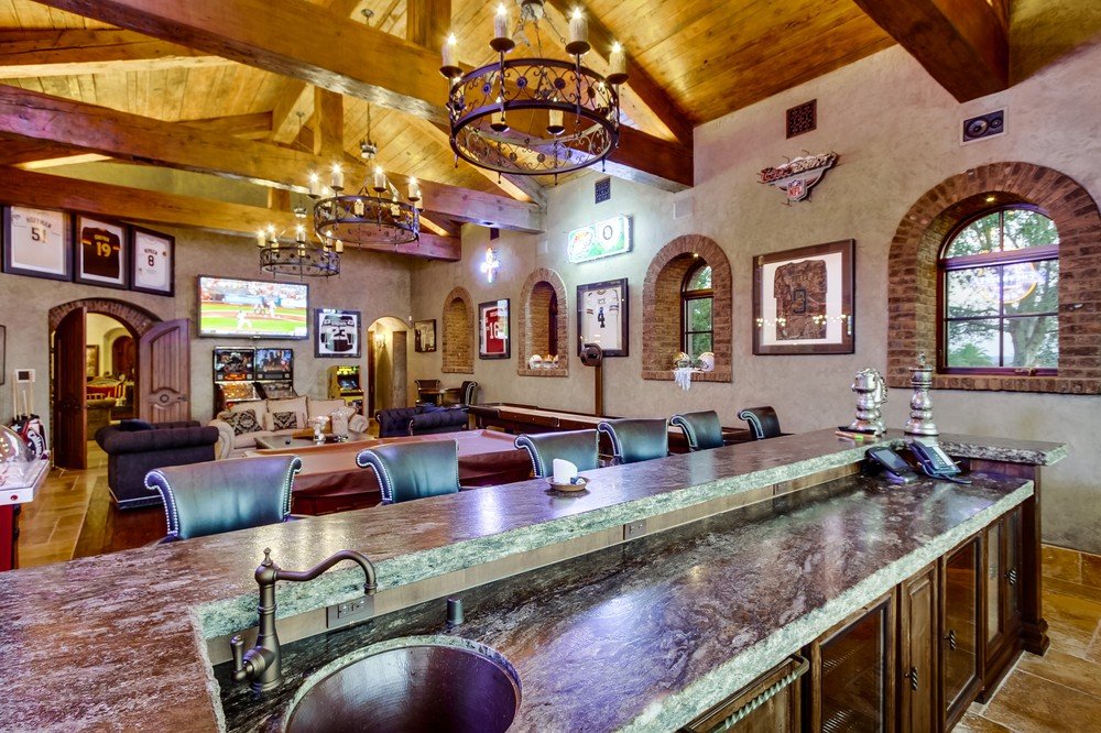 Indoor lounge area with a bar counter in the foreground, high-backed chairs, chandeliers hanging from a wooden ceiling, sports on multiple TVs, framed pictures, windows with brick arches, and cozy seating in the background.
