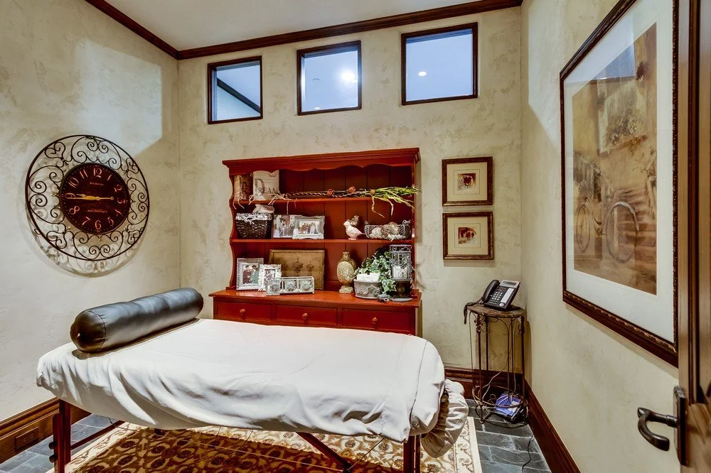A massage room with a massage table, decorative wall clock, wooden cabinet with photo frames and decorative items, framed artwork, and a small side table with a landline phone.