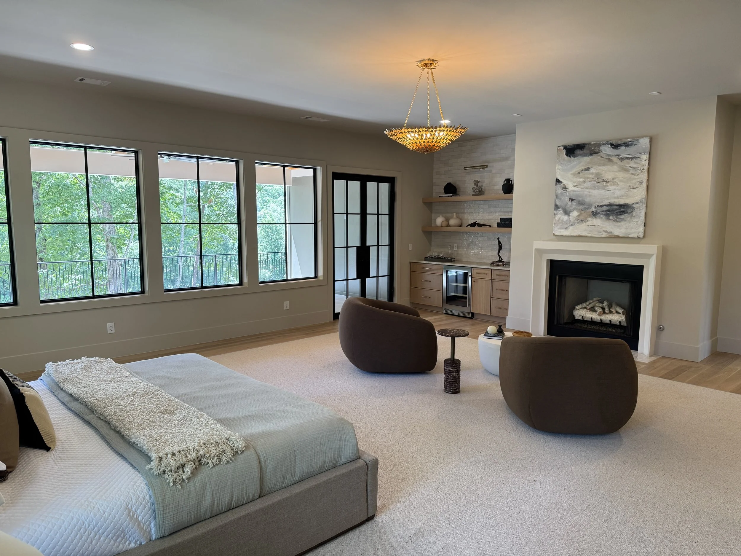The image shows a modern living room with large windows, a fireplace with a mirror above it, and a ceiling light fixture. There are two armchairs, a small table, and a bed or sofa with blankets and pillows.