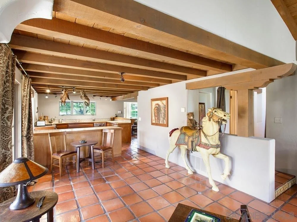 Living room with wooden beams on the ceiling, terracotta tiled floor, a wooden table with chairs, and several horse and animal decorations, including a model of a horse with a saddle and a bear on a ledge.