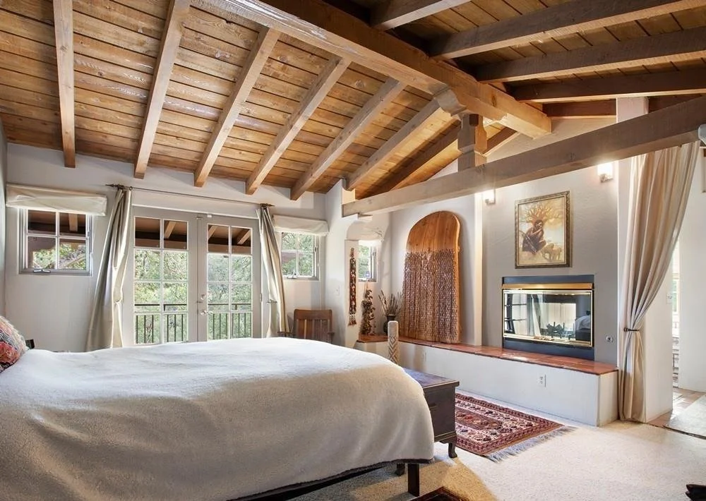 A bedroom with a vaulted wooden ceiling, large windows, and glass doors leading to a balcony. The room features a bed, a small wooden chair, a tapestry on the wall, and a fireplace built into the wall with a wooden mantel. Light-colored curtains fram