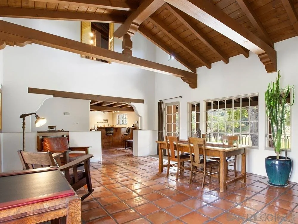 Room with wooden ceiling beams, tiled floor, large windows, and a wooden dining table with chairs. There is a tall potted plant near the windows and a small seating area with chairs and a side table.