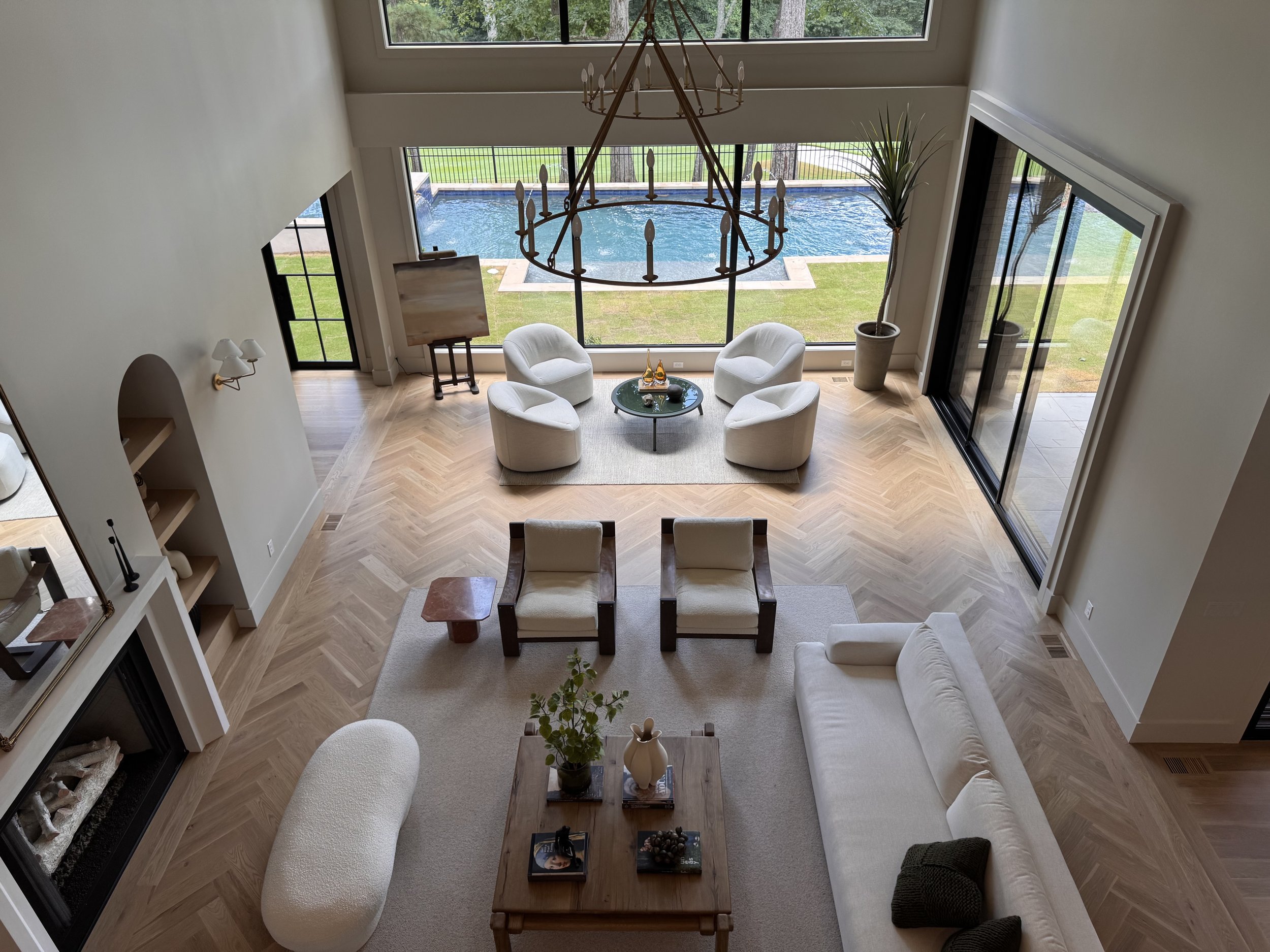 Living room with white furniture, a glass coffee table with books and decorative items, a large window showcase overlooking a pool outdoor, tall potted plants, and a chandelier hanging from the ceiling.