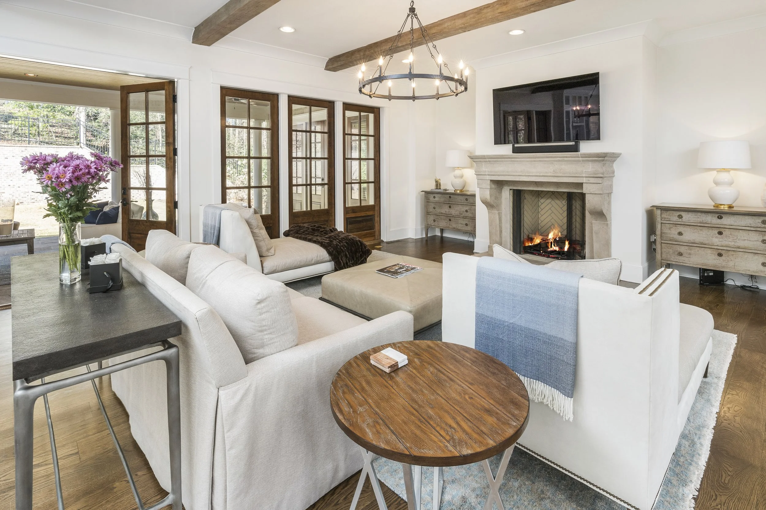 Living room with beige sofas, a round wooden coffee table, a fireplace, a large flat-screen TV above, and glass doors leading outside. There are decorative lamps, a vase of pink flowers, and a rustic chandelier hanging from the ceiling.