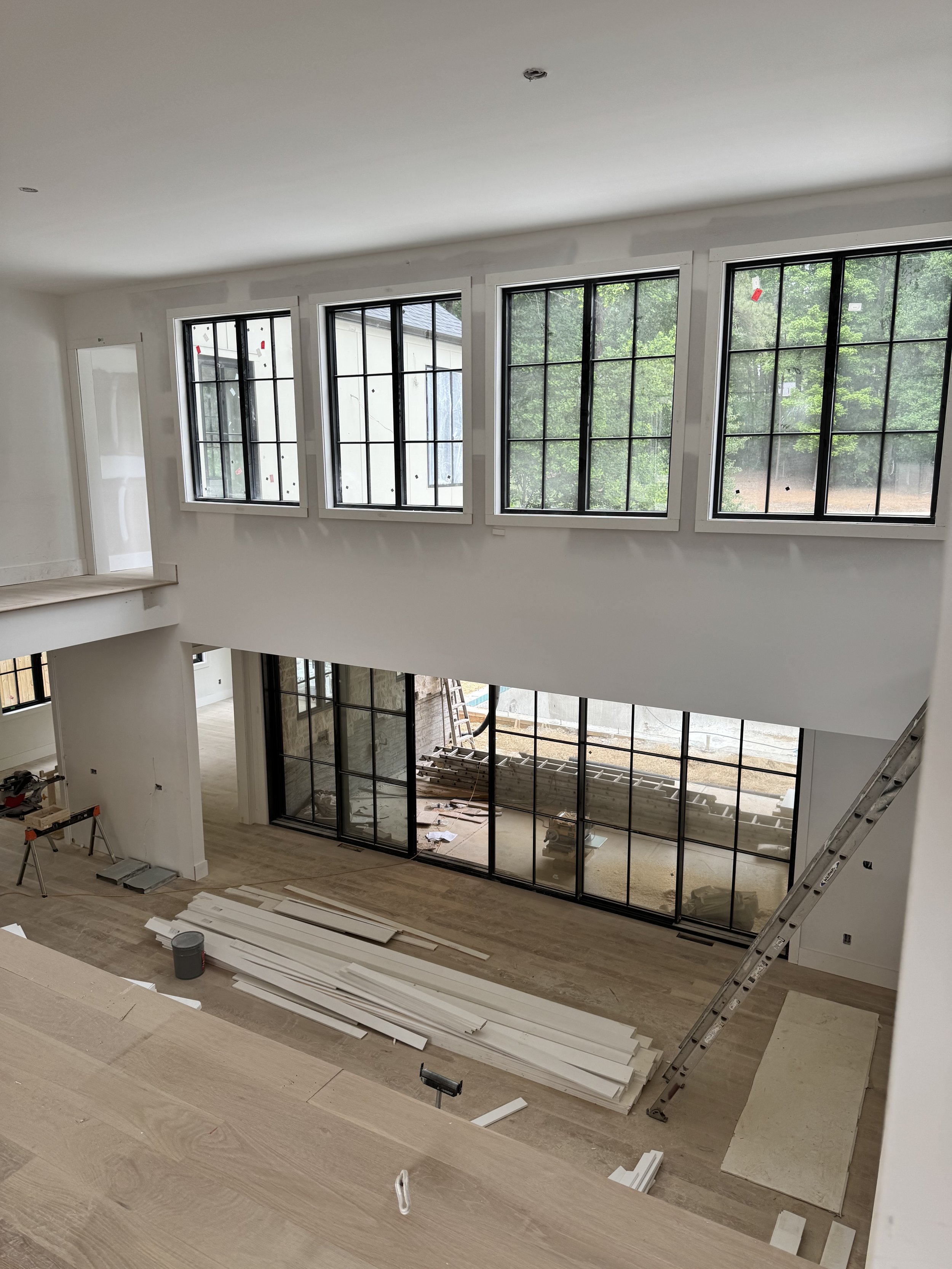 Interior of a house under construction with large black-framed windows, unfinished walls, and construction materials on the floor.
