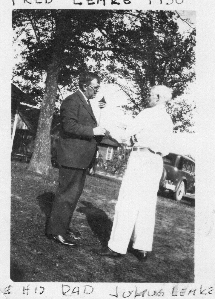 A black-and-white photograph of two men shaking hands outdoors, with trees and a vintage car in the background. The older man has white hair and is wearing light-colored pants and a shirt, while the younger man wears glasses and a dark suit. Handwritten text is visible above and below the image, indicating it might be a personal or family photograph.