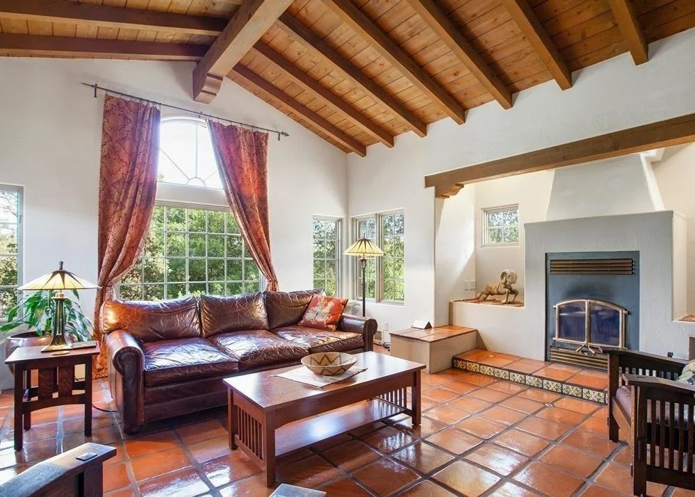 Living room with large window, leather sofa, wooden coffee table, tiled floor, wooden ceiling, fireplace, and decorative items.