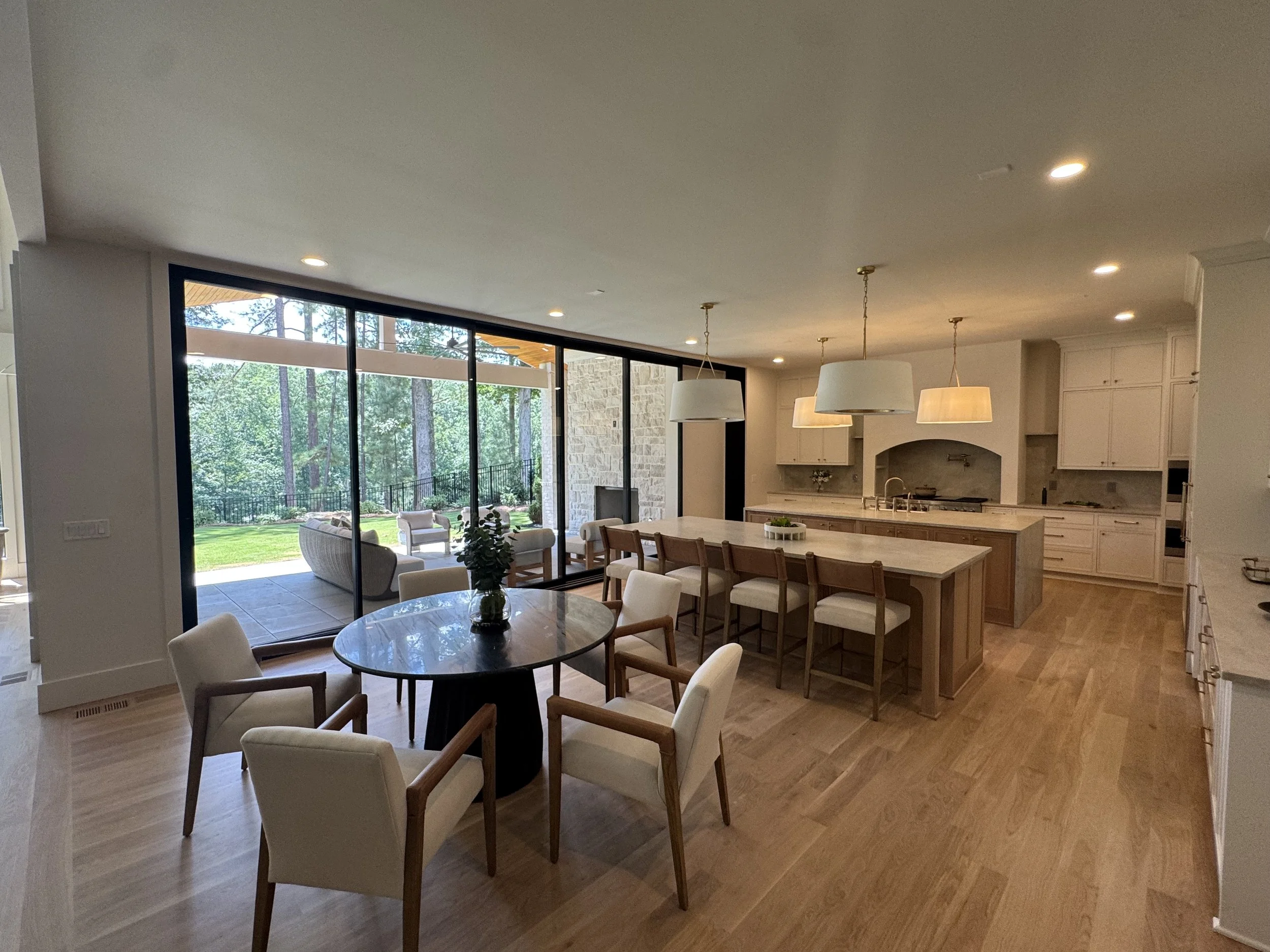 Open living space with large windows showing trees outside, a dining area with a round table and white chairs, and a kitchen with a white island and pendant lights.
