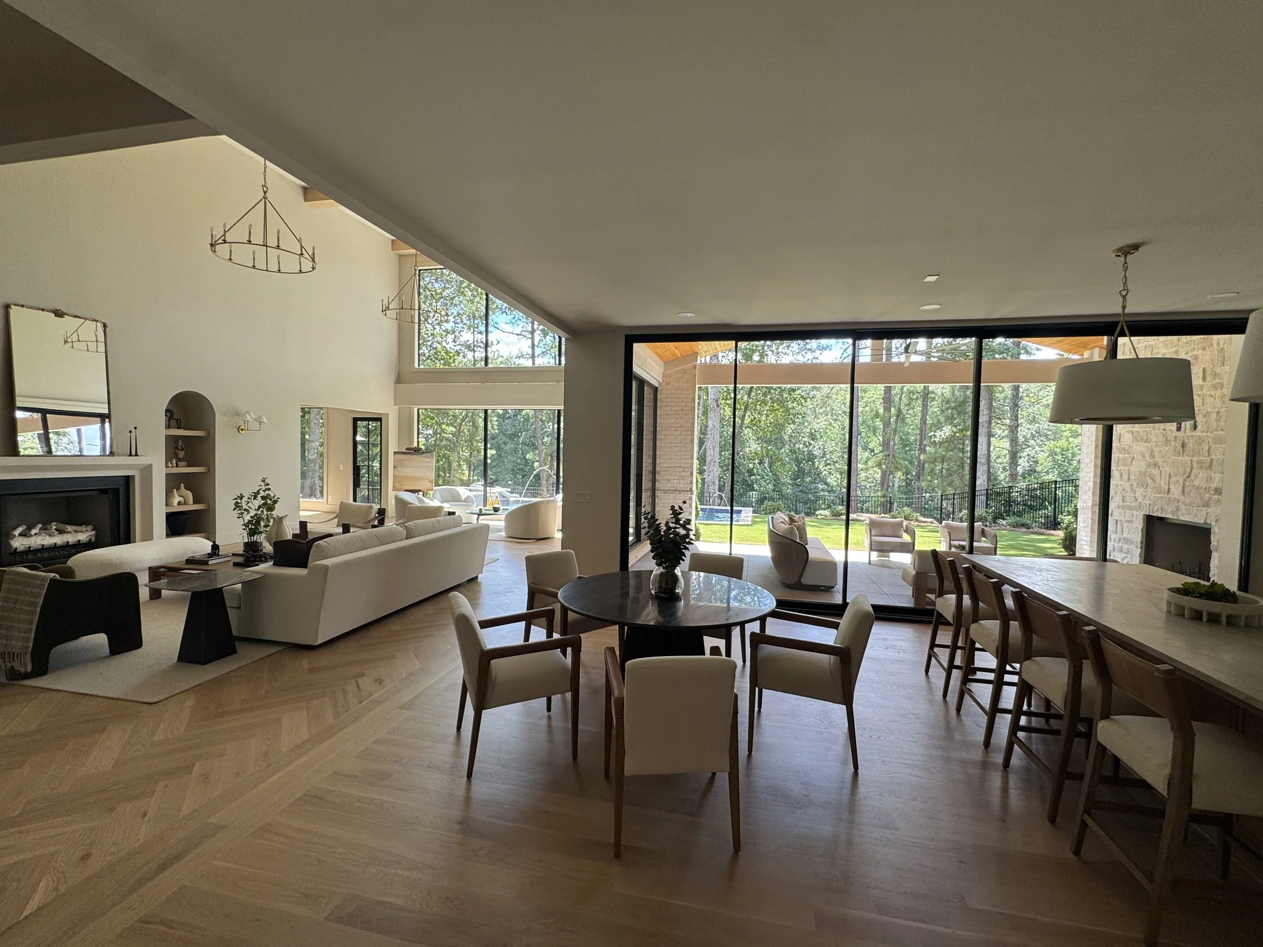 Spacious modern living room with large floor-to-ceiling windows providing views of a green outdoor area, featuring white sofas, a round dining table with chairs, and minimalistic decor.