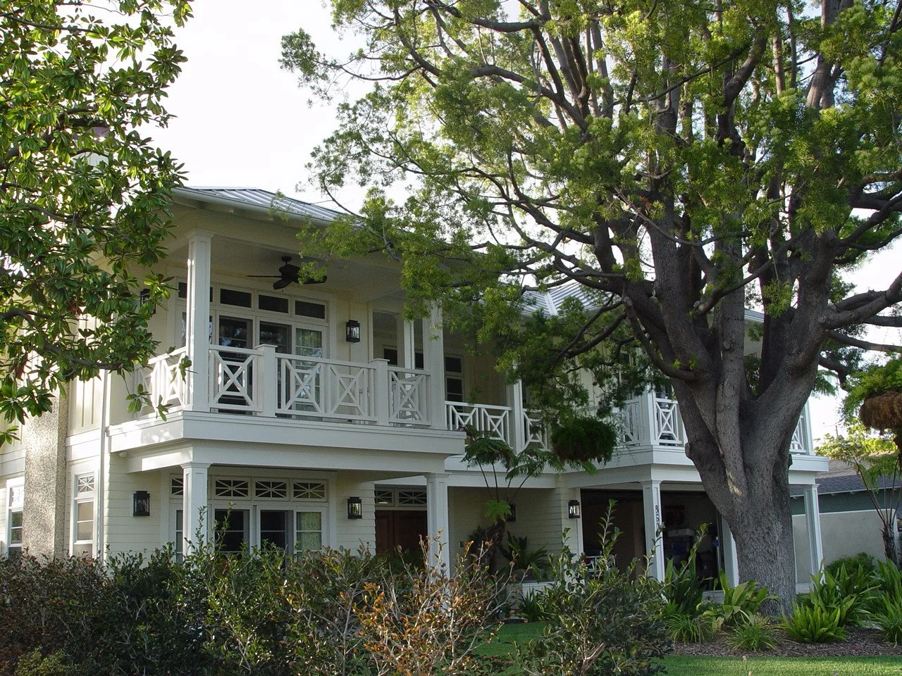 A two-story house with a large front porch, white siding, and a balcony on the second floor surrounded by trees and greenery.