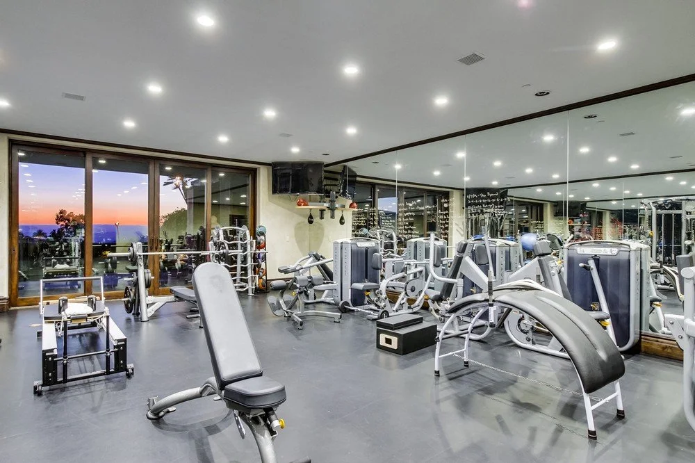 Gym room with workout equipment including treadmills, weight machines, and free weights, with a large mirror wall and windows showing sunset outside.