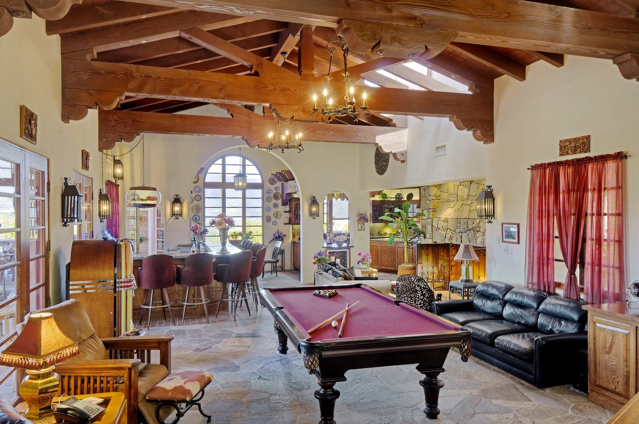 Interior of a living room with wooden beams on the ceiling, chandelier, large window, fireplace, and various furniture including chairs, sofas, and a pool table.