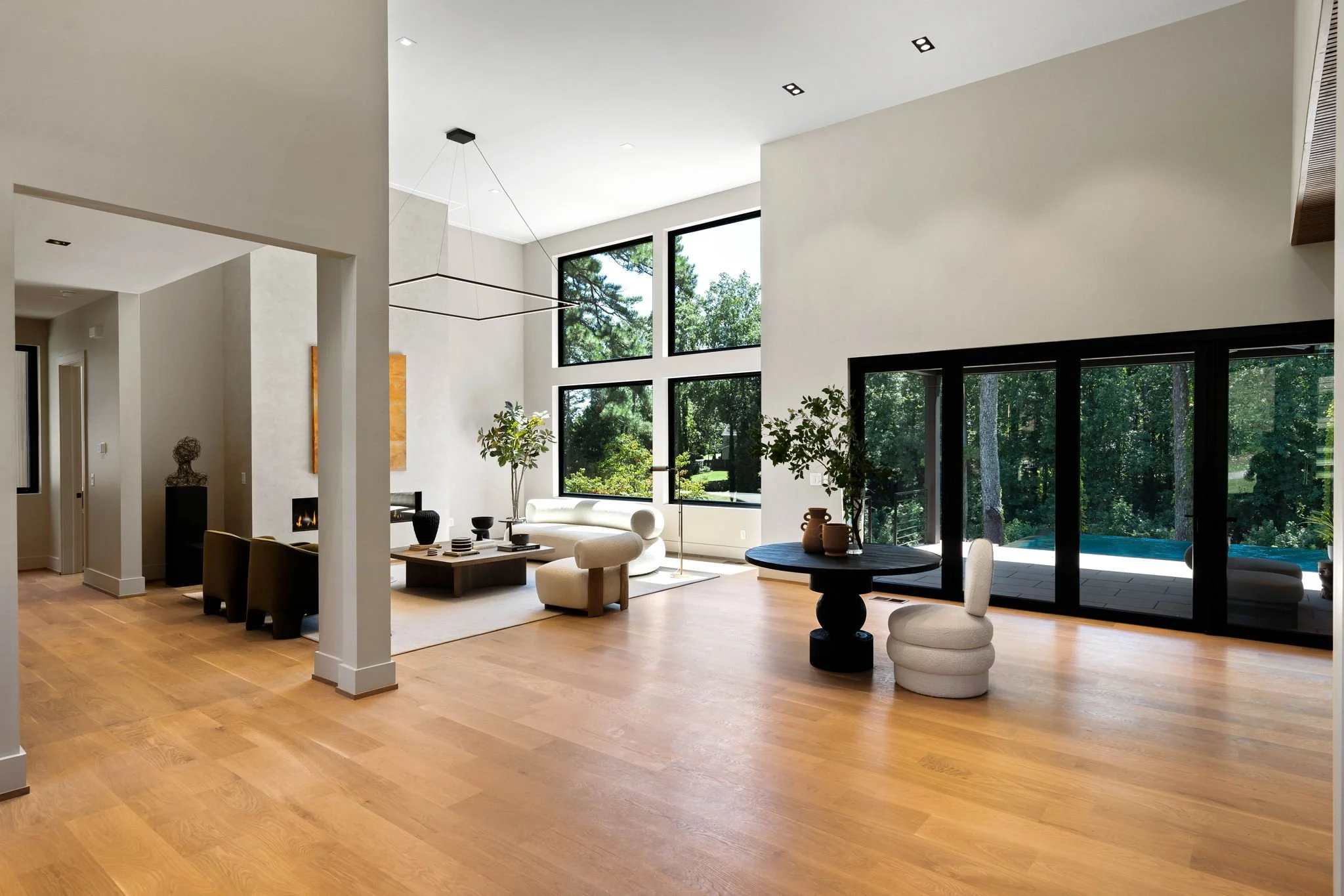 Modern living room with large windows, white walls, hardwood flooring, contemporary furniture, indoor plants, and access to outdoor patio with swimming pool.