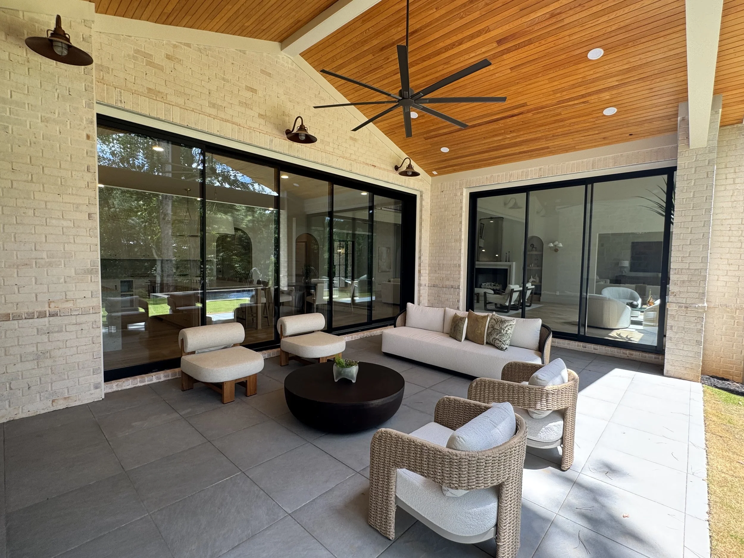 Covered outdoor patio with beige brick walls, large windows, and a wooden ceiling. The patio features outdoor furniture including a white sofa, woven chairs, and a black round table. A ceiling fan hangs from the ceiling, and sunlight streams in throu