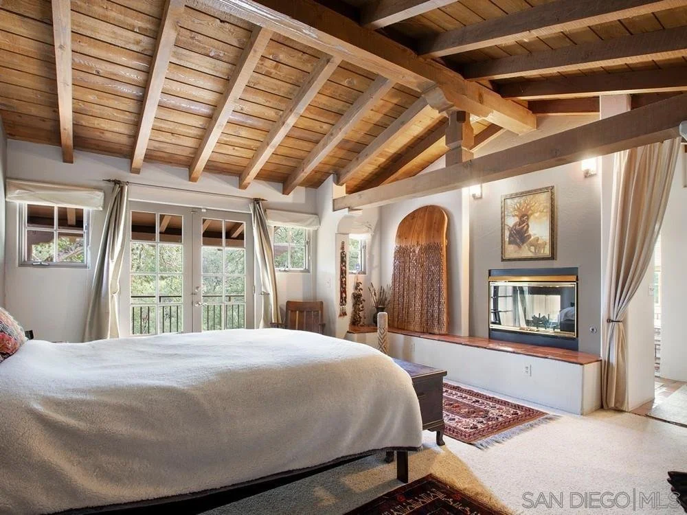 Bedroom with vaulted wooden ceiling, large bed with white bedding, multiple windows with curtains, a fireplace on the wall, and a small balcony door.