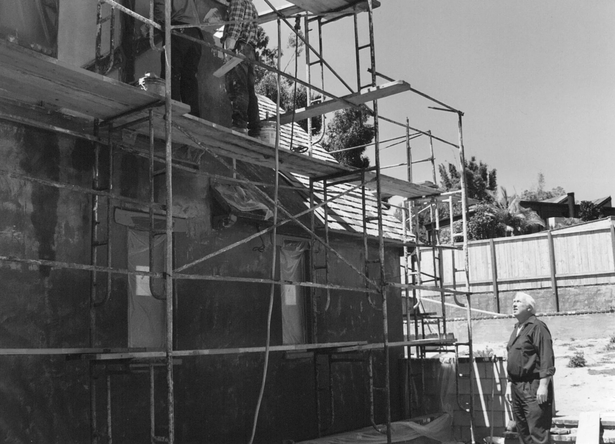 Black and white photo of construction workers working on a home renovation. One worker is on scaffolding, and another is standing on the ground beside the building with a tool in hand. The house has a partially installed roof and a fenced backyard with trees in the background.
