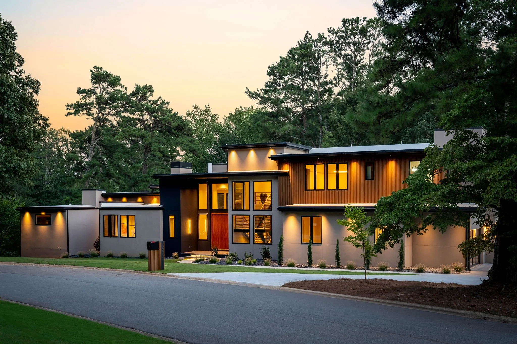 Modern multi-story house with illuminated windows surrounded by tall trees at dusk.