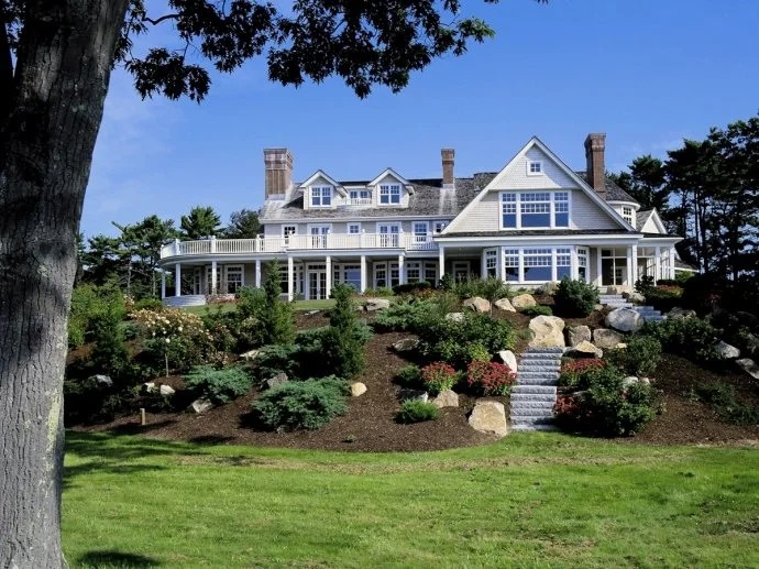 Large white house on a hill with multiple stories, surrounded by landscaped garden and stone steps, with a large tree in the foreground.