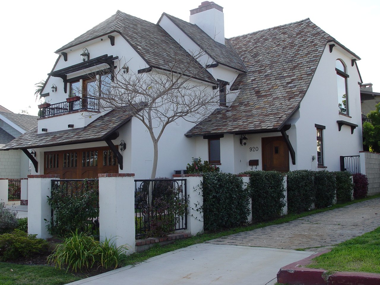 White two-story house with brown shingle roof, small front yard with bushes, tree, and black metal fence, located in a suburban neighborhood.