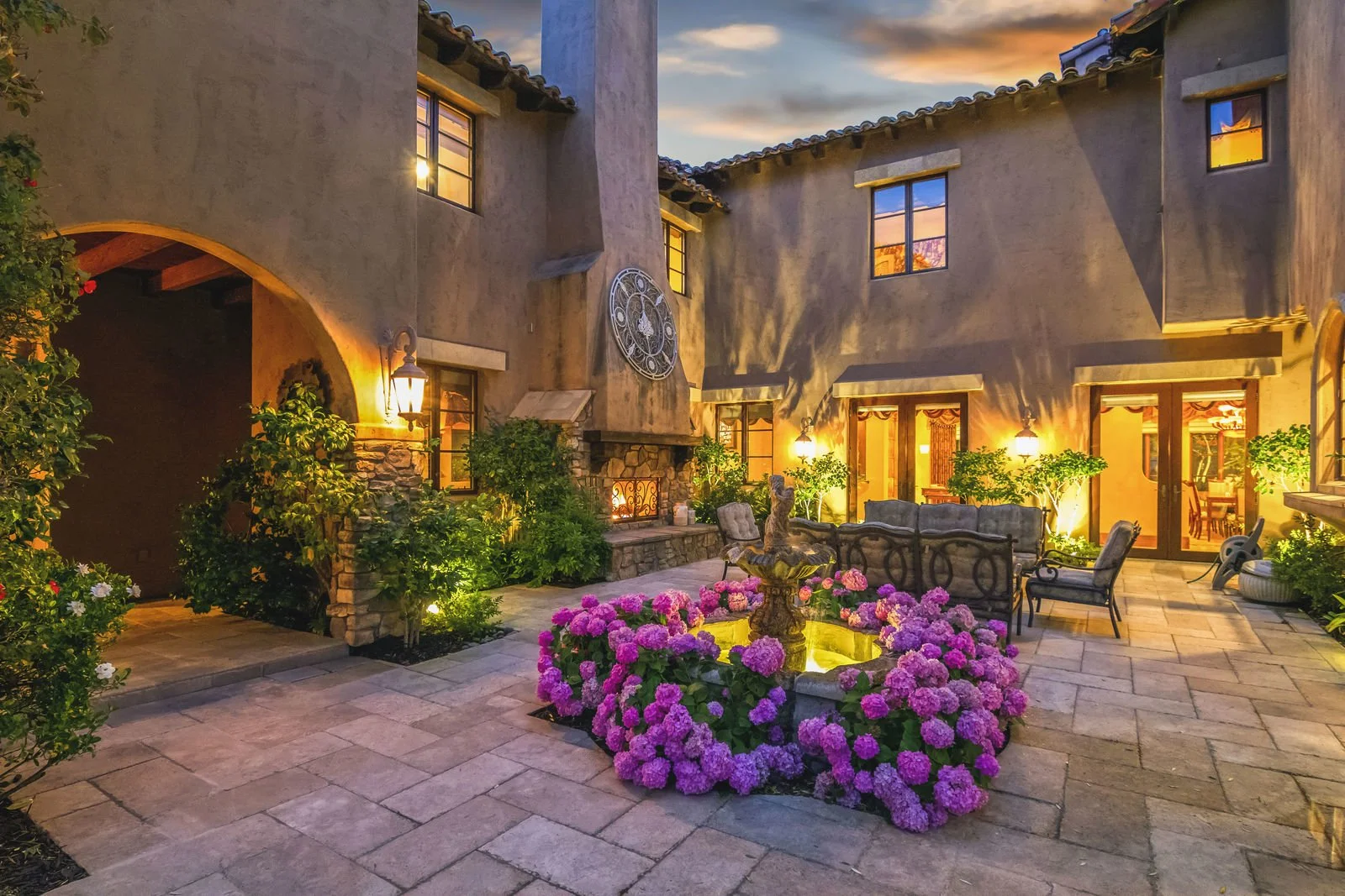 A cozy courtyard of a house at dusk, illuminated with warm lighting. There is a central fountain surrounded by pink hydrangeas, outdoor seating, and lush green plants. The house features stone and stucco walls with multiple windows and glass doors.