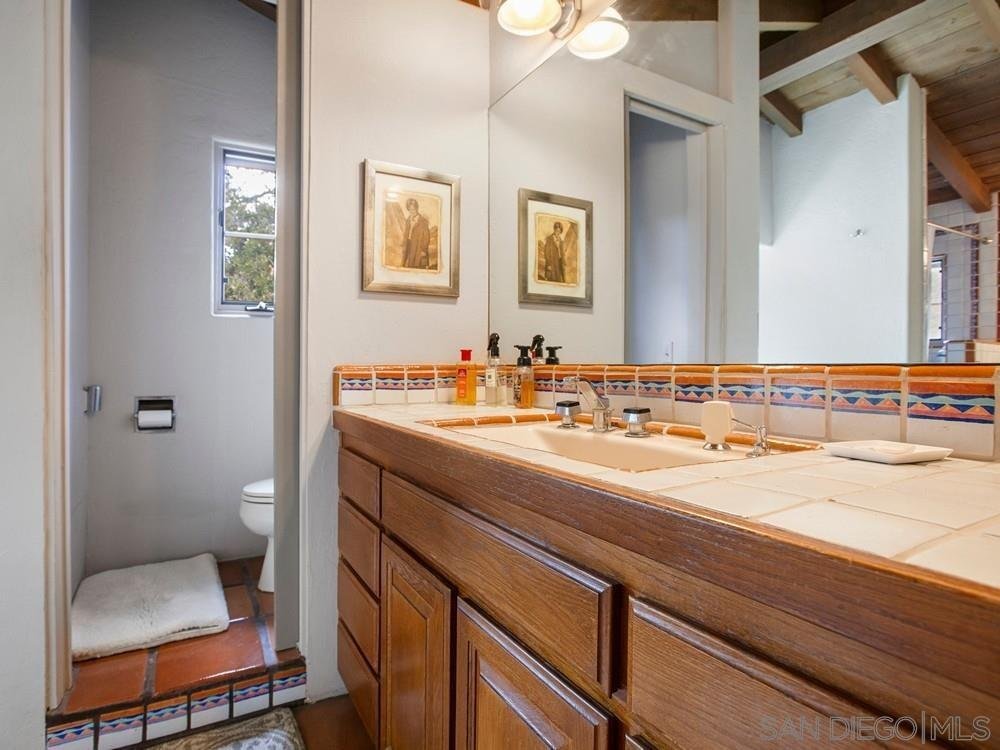 Bathroom with wooden vanity, large mirror, framed artwork, and a separate toilet area with window and small shelf.