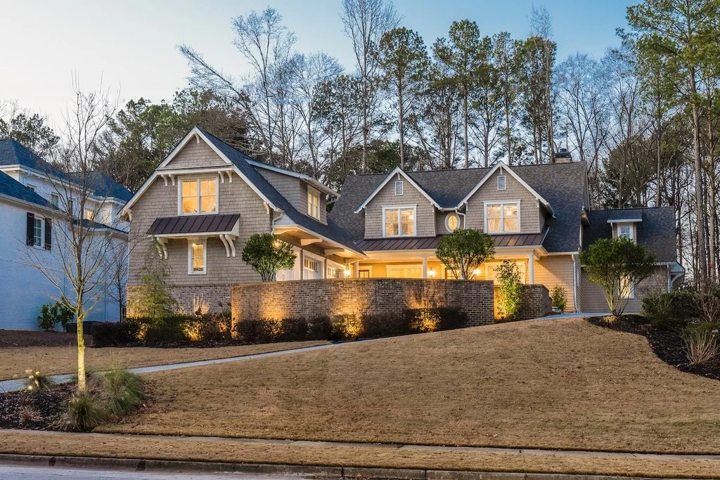 Exterior view of a large, two-story house with beige siding, multiple gabled roofs, and porch lighting, surrounded by a landscaped yard with grass, trees, and shrubs at dusk.