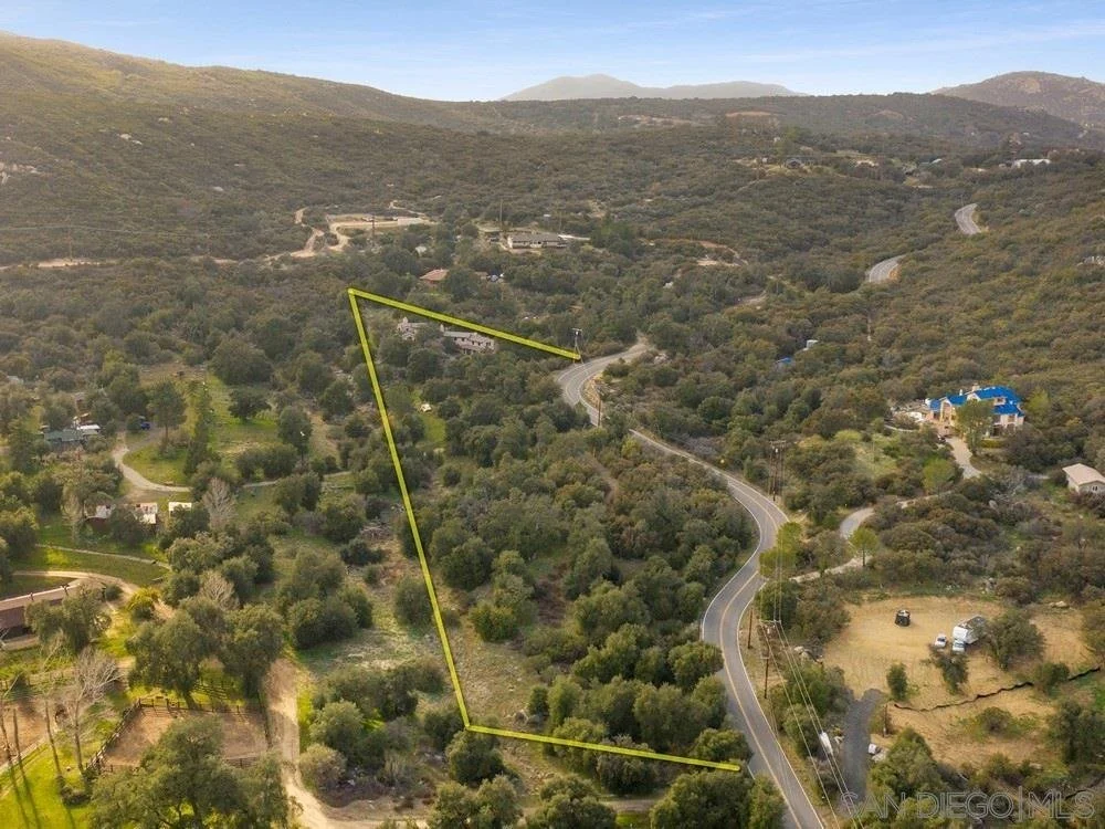 Aerial view of a curvy road running through a hilly, forested area with a few houses and open spaces, with one property outlined in yellow.