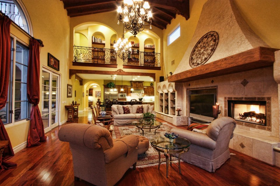 Spacious living room with high ceilings, large windows, a fireplace, chandelier, wooden beams, and comfortable furniture including a sofa, armchair, and glass coffee tables.