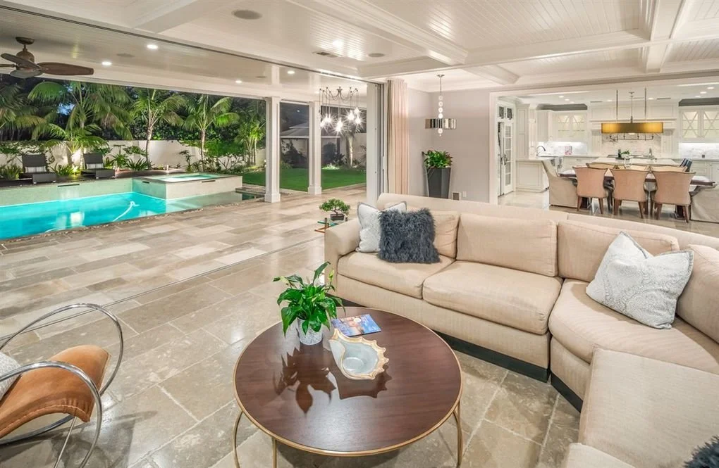 Open-concept living room with beige sectional sofa, coffee table with a plant and decorative tray, overlooking a backyard with a swimming pool and lush greenery, adjacent to a dining and kitchen area.
