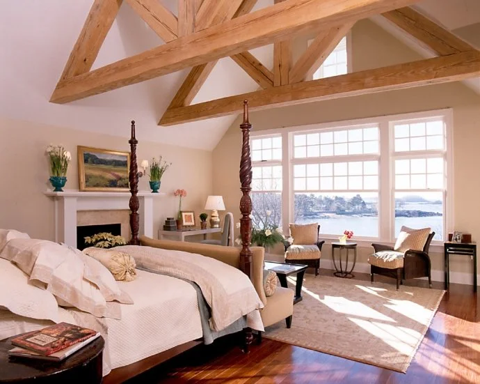 Bright bedroom with vaulted ceiling, large window overlooking water, four-poster bed, seating area with two chairs, side tables, and decorative plants.