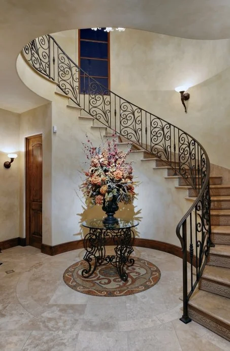 Indoor curved staircase with ornate iron railing, a decorative floral arrangement on a table, and two wall-mounted light fixtures.